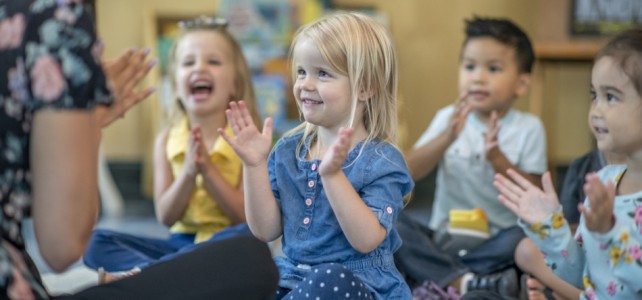 A small group of multi-ethnic preschool children sit on the floor in front of their teacher as she teaches them a song.  They are each dressed casually and sitting with their legs crossed as they clap and sign along.